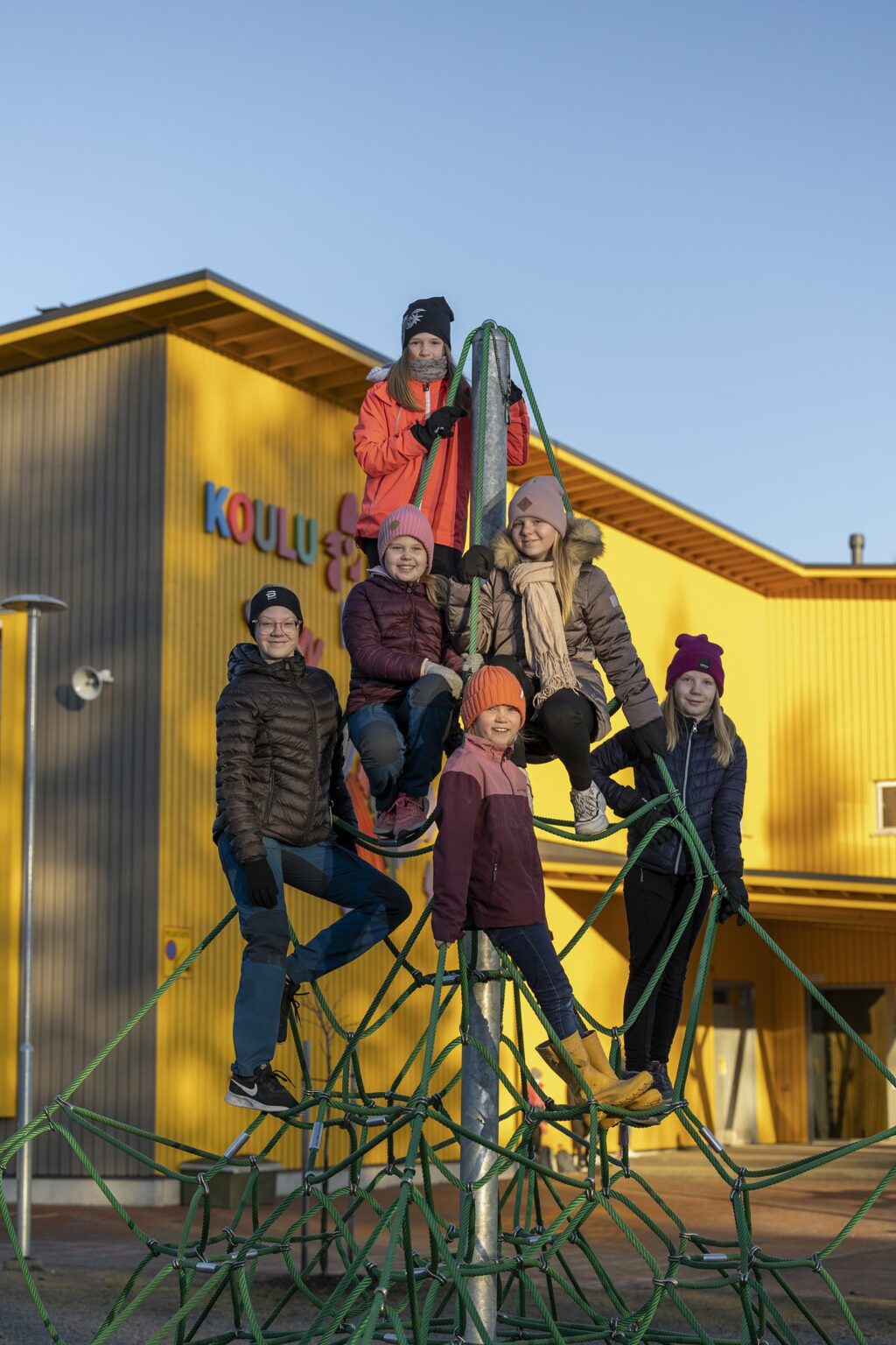 Kuusi lasta on kiivennyt kiipeilypyramidiin ja katsovat kohti kameraa iloisina. Osa lapsista istuu ja osa seisoo köysillä.