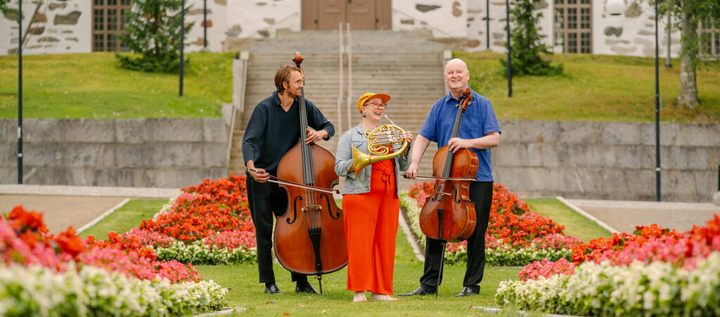 Kolme orkesterin muusikkoa soittimien kanssa puistossa.