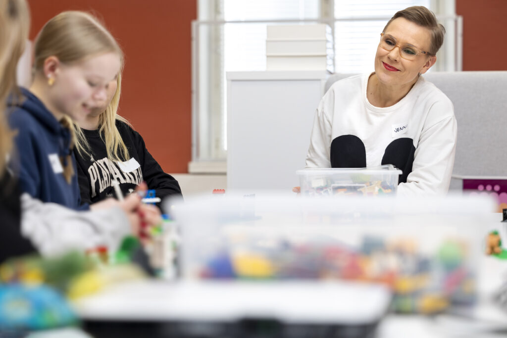 Nuoria rakentamssa legoja, työpajan ohjaaja katsoo hymyillen.
