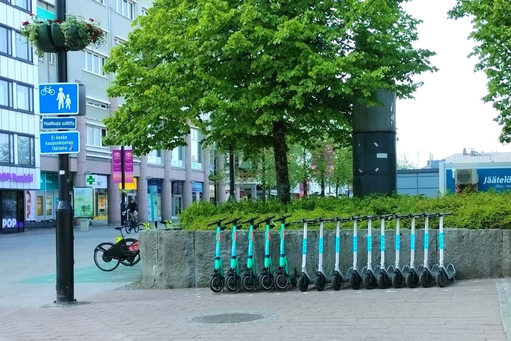 Sähköpotkulautoja pysäköitynä siistissä rivissä kivimuurin edessä lehmuspuiden alla.