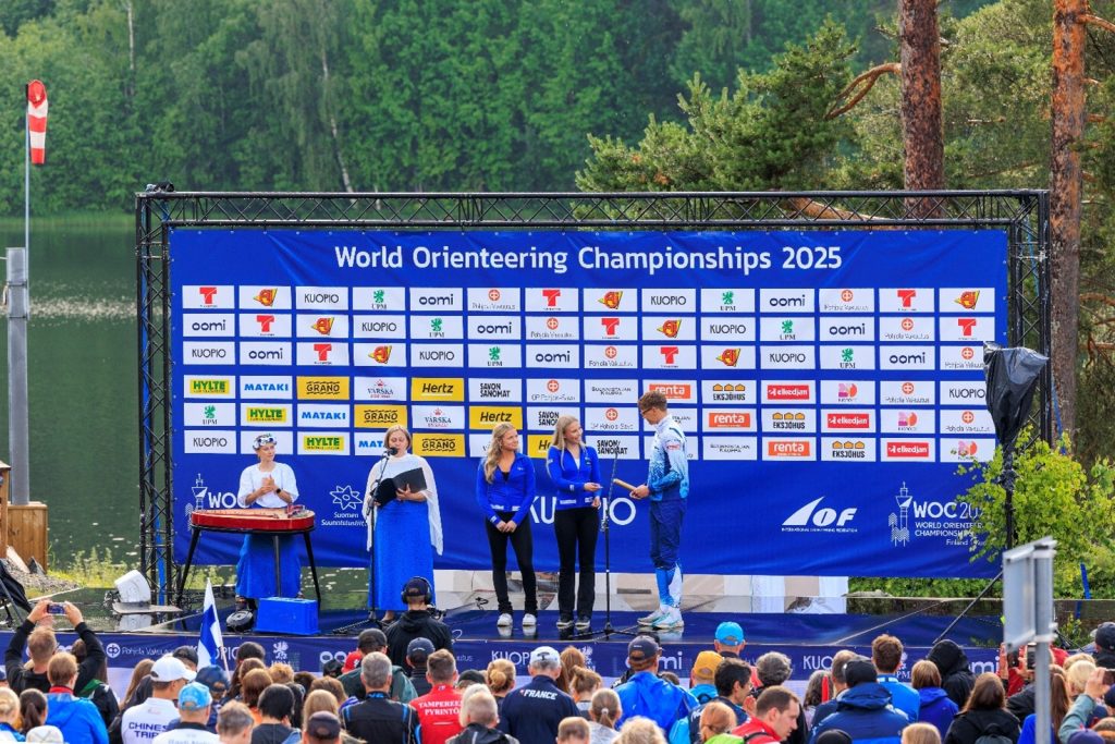 WOC2025 tapahtumalavalla kaksi taitoluistelijaa antaa kultaisen viestikapulan suunnistajalle