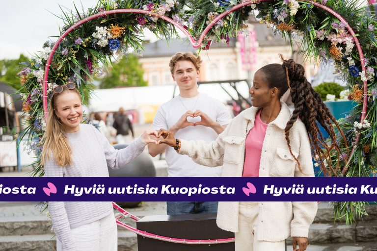 Kolme nuorta nauraa sydämenmuotoisen kukkaköynnöksen keskellä.