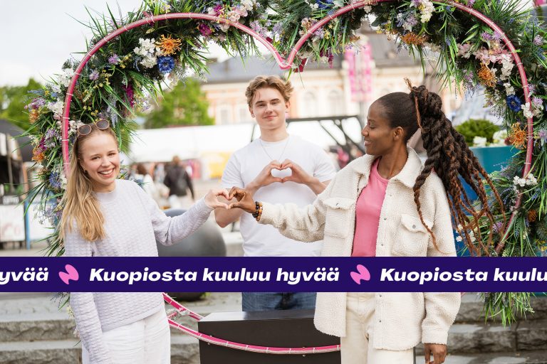 Kolme nuorta nauraa sydämenmuotoisen kukkaköynnöksen keskellä.