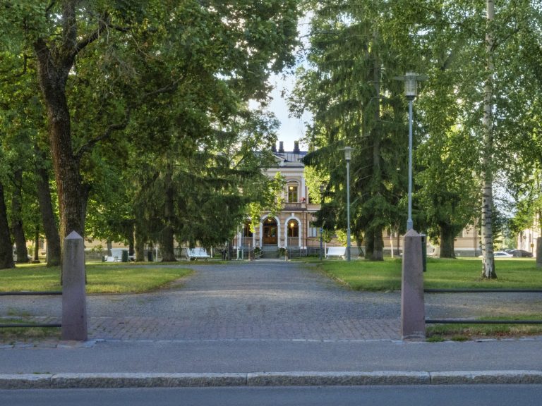 Puistomainen alue, jossa on tiilillä päällystetty kävelytie. Tien molemmilla puolilla on kiviset pylväät, jotka muodostavat porttimaisen sisäänkäynnin. Taustalla kohoaa suuri ja koristeellinen rakennus. Rakennuksessa on kaari-ikkunoita ja koristeellisia yksityiskohtia julkisivussa. Alueella on myös hyvin hoidettu nurmikko ja kävelytien varrella lamppuja.