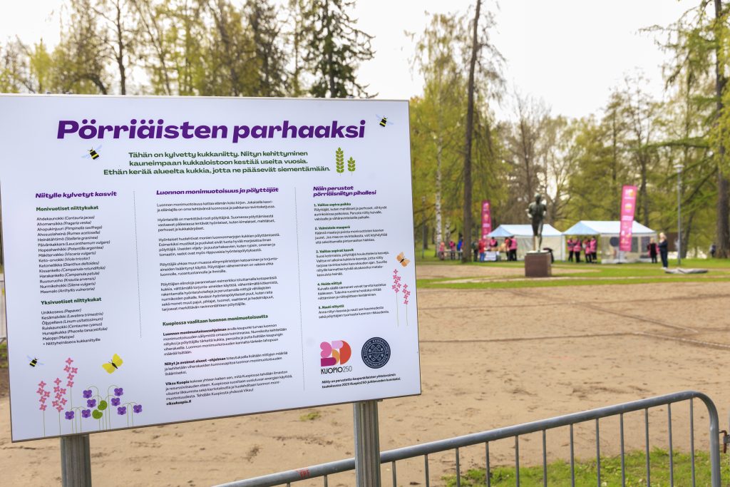 Suuri opastaulu ulkona puistossa, otsikossa lukee ”Pörriäisten parhaaksi”. Taulussa on tietoa niittyjen hyödyistä, luonnon monimuotoisuuden lisäämisestä ja ohjeita pölyttäjien auttamiseen. Taulun alareunassa näkyy Kuopion kaupungin logo. Taustalla on hiekkakenttä, puita ja telttakatoksia, joissa on ihmisiä sekä pinkkejä tapahtumalippuja.