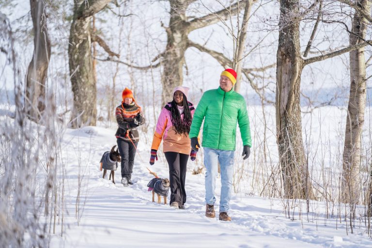 Talvisessa, kirkkaassa päivän valossa kolme kävelijää puiden lomassa. Mukana on myös kaksi koiraa.