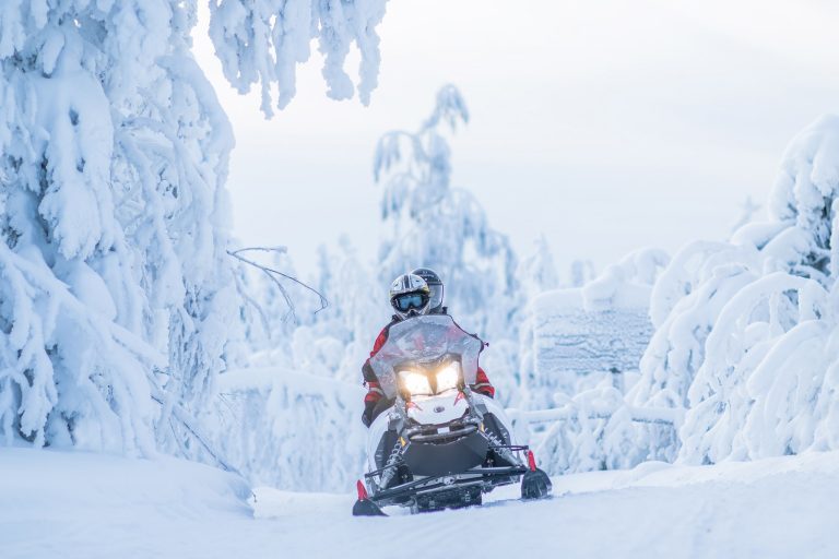 Talvinen maisema, lumisisa puita taustalla. Moottorikelkka tulee kohti, jonka kyydissä kaksi henkilöä.