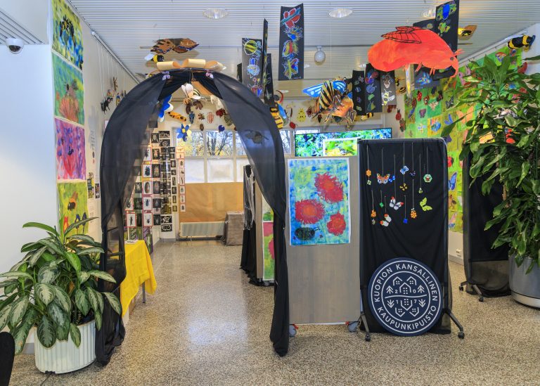 Sisätila, jossa on värikäs näyttely. Seinillä ja katossa roikkuu maalauksia ja askarteluja, kuten kukka-aiheisia tauluja ja perhoskoristeita. Keskellä kulkuaukko, jossa musta kangaskehys. Näyttelytilassa on myös suuria viherkasveja ja Kuopion kansalaisopiston tunnuksella varustettu musta kangas.