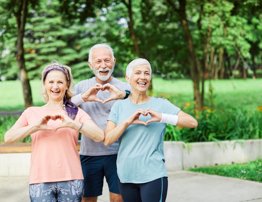 Kaksi nauravaa ikääntynyttä naista ja yksi naurava ikääntynyt mies seisovat ryhmänä ja tekevät käsillä sydämen muodon. Taustalla vihreä niitty ja puita.