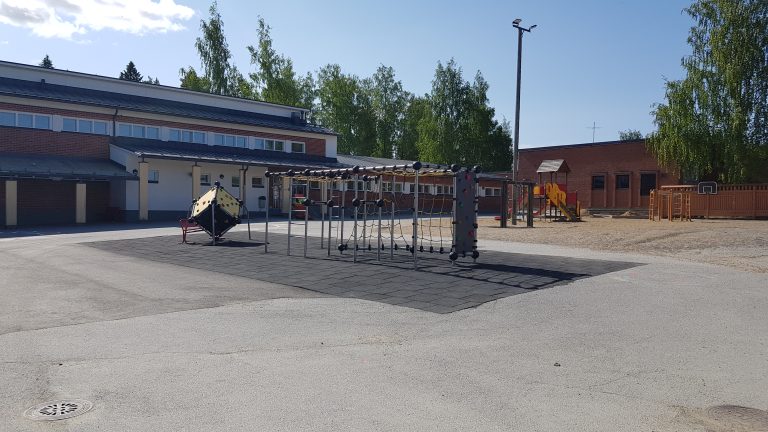 Asfaltoidulla pihalla leikkivälineitä, taustalla punatiilinen rakennus.