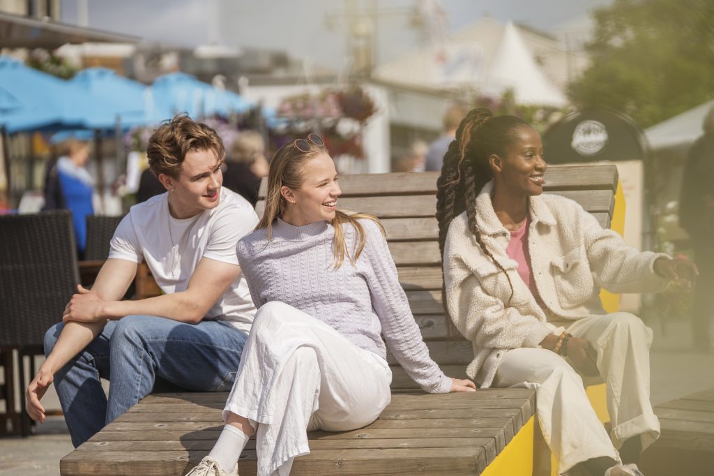 Kolme henkilöä istuu kesäisenä päivänä puurakentaisella tuolilla.