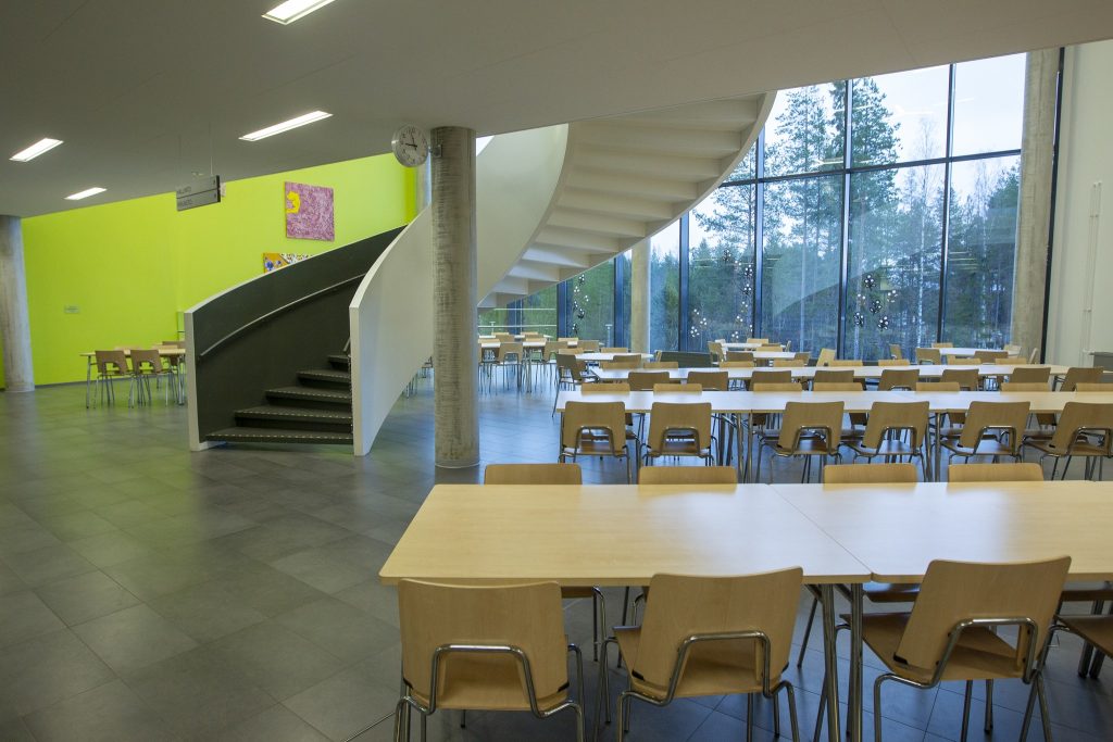 Avara koulun ruokala, jossa on riveittäin vaaleita pöytiä ja tuoleja, suuret ikkunat metsään sekä keskellä tilaa kaartuvat portaat.