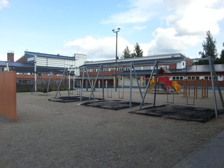 uva koulun pihasta, jossa on useita metallisia keinutelineitä hiekkakentällä. Taustalla näkyy matalia koulurakennuksia ja leikkipaikan kiipeilytelineitä. Taivas on osittain pilvinen.”