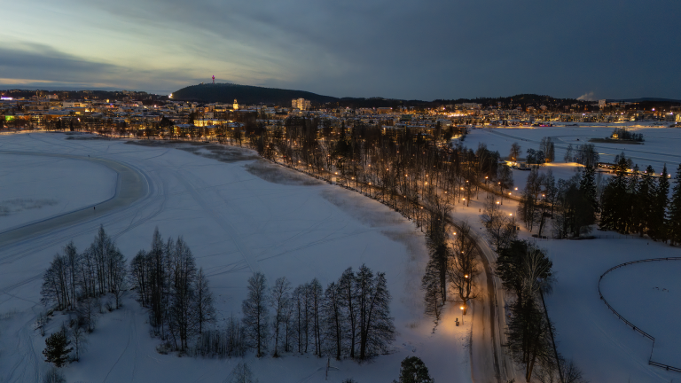 Talvinen iltanäkymä kaupungista: etualalla on luminen puistoalue ja jäinen vesialue, jonka reunalla kulkee valaistu kävelytie puiden lomassa. Taustalla näkyy laaja kaupunkimaisema talojen ja katuvalojen loisteessa hämärtyvää taivasta vasten.