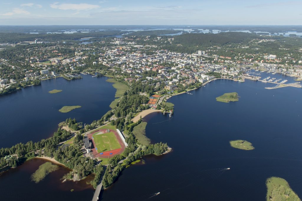 Ylhäältä päin kuvattuna vehreä Väinölänniemi, jossa urheilukenttä. Kallavettä ja taustalla kaupungin keskustan rakennuksia ja Puijon alue.