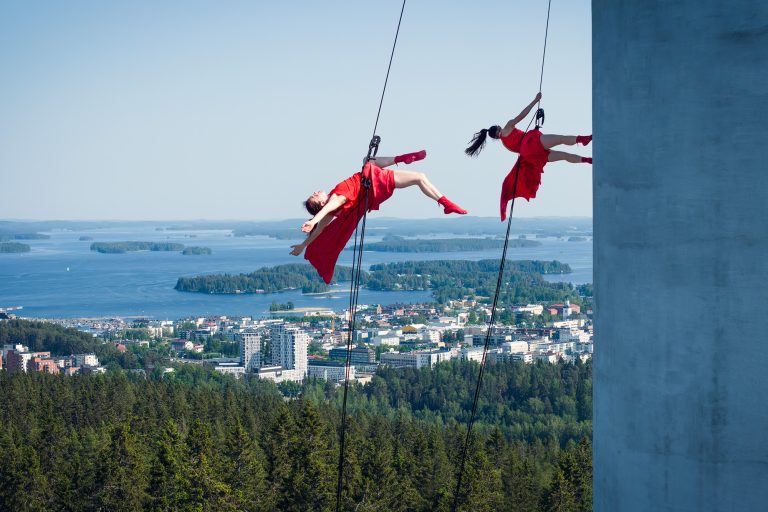 Kaksi punapukuista naista tanssii ilmassa Puijon tornin kyljessä.