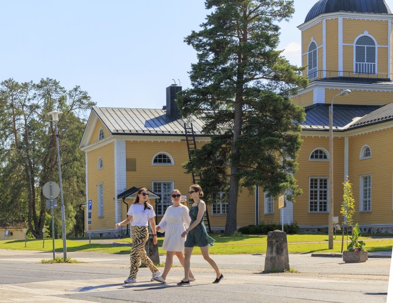 Kolme naista kävelee kesäisessä Maaningan keskustassa, keltaisen puukirkon edustalla.