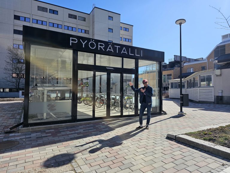 Kuopion pyörätalli aurinkoisessa säässä, etualalla hymyileväinen henkilö tallin edustalla.