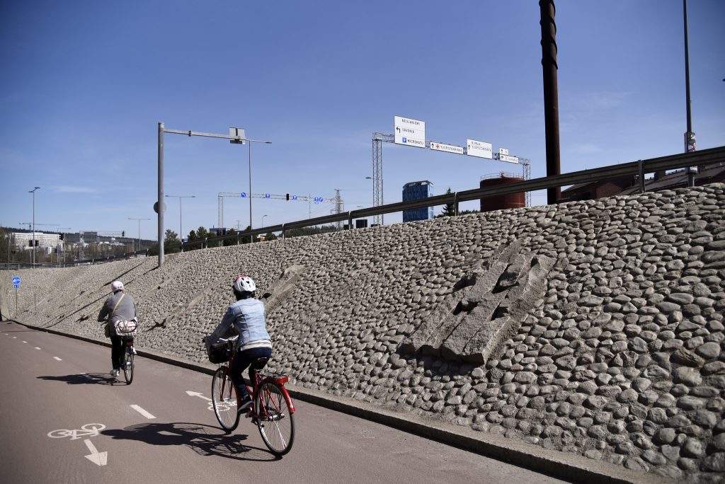 Harmaalla kiveyksellä reunustetulla pyörätiellä ajaa kaksi pyöräilijää, taustalla sinininen taivas.