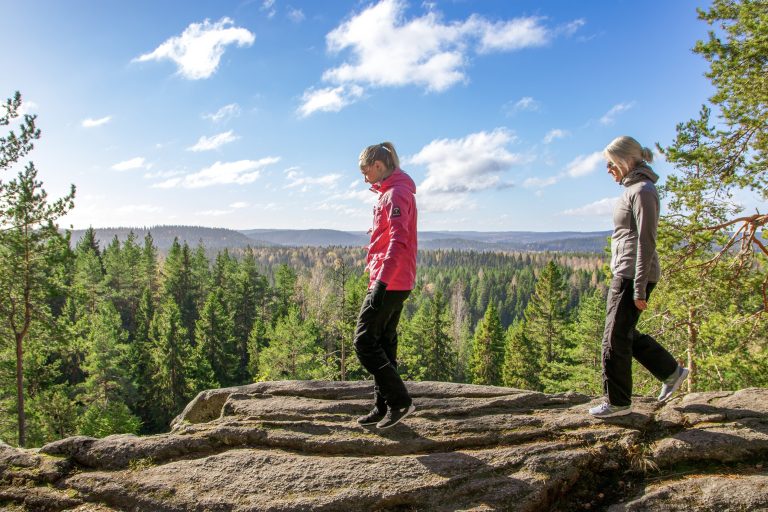 Kaksi naishenkilöä kävelee kalliolla, taustalla puita ja sininen taivas.