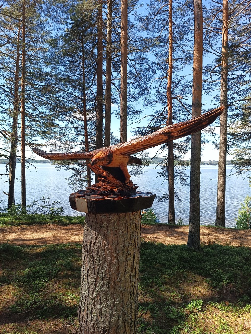 Kalasääskiveistos katkaistun puunruongon päällä, taustalla järvimaisemaa.