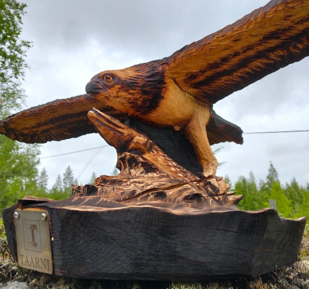 Ruskea, puinen veistos, joka esittää kalasääkseä. Taustalla pilvinen taivas ja metsää.