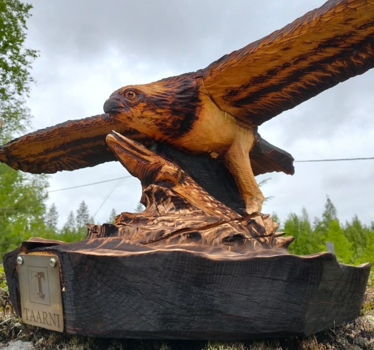 Ruskea, puinen veistos, joka esittää kalasääkseä. Taustalla pilvinen taivas ja metsää.