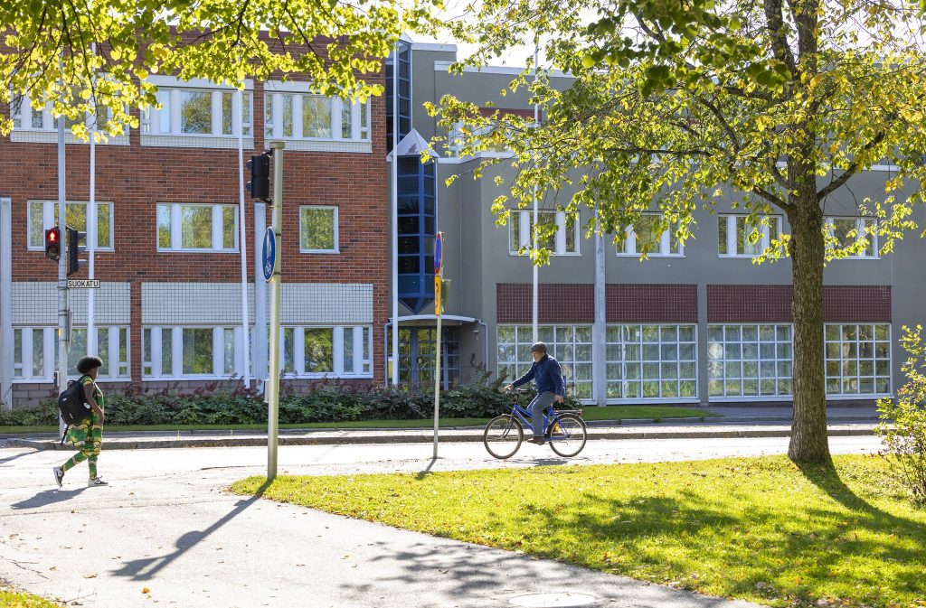 Punavalkoinen rakennus, jonka edustalla vihreitä puita ja pyöräilijä ja jalankulkija.