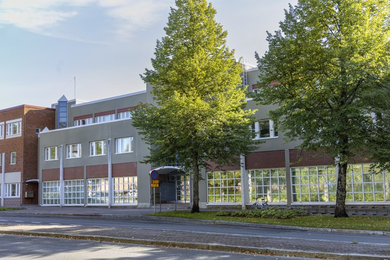 Punaharmaa rakennus, jonka edustalla kaksi vihreää puuta. Rakennuksen edessä katu ja taustalla sininen taivas.