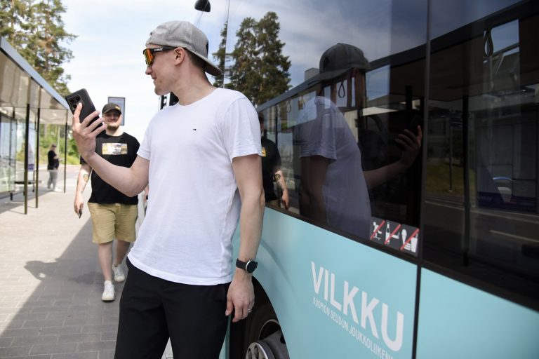 Bussin vieressä seisoo kaksi mieshenkilöä. Toinen miehistä katsoo matkapuhelintaan.