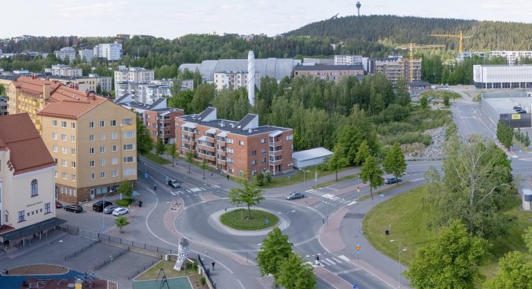 Liikenneympyrä, sitä ympäröiviä kerrostaloja, katuja, pyöräteitä ja viheralueita. Taustalla näkyy metsää, rakennuksia sekä Puijon torni.