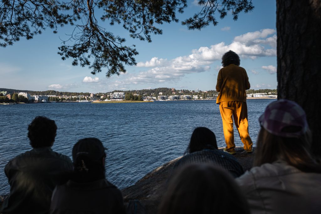 Järven rannalla kallion päällä seisova henkilö katsoo kohti vastarannan kaupunkimaisemaa. Etualalla useita katsojia istuu tai seisoo puiden varjostamassa rinteessä. Taustalla näkyvät sininen järvi, pilviä täynnä oleva taivas sekä rantaviivaa seuraava kaupunkinäkymä.