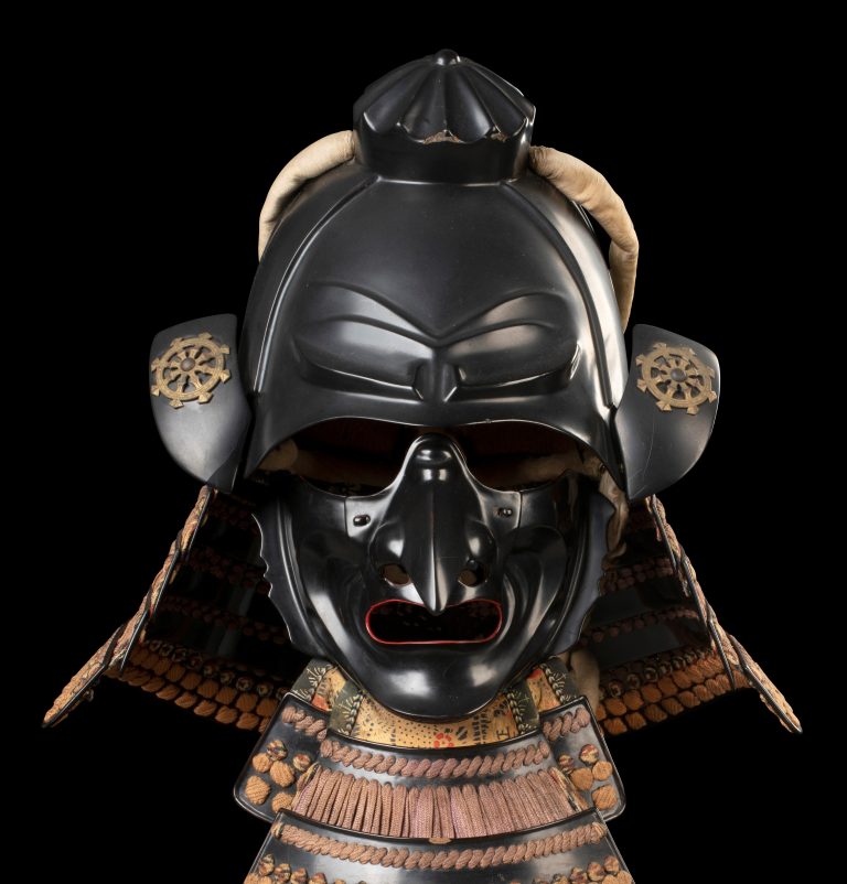 Front-facing Japanese samurai helmet and armor in black lacquer with gold details, displayed against a black background. The ornate armor features decorative cords, layered neck protection, and a fierce face mask.