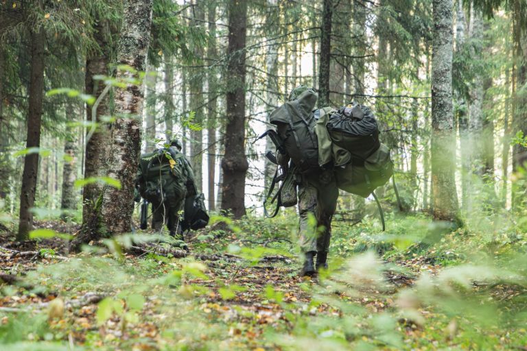 Kaksi suuria kantamuksia kantavaa varushenkilöä kävelee havumetsässä metsäpolkua pitkin. Ympärillä on tiheää metsää, sammalta ja aluskasvillisuutta.