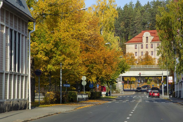 Syksyinen Maaherrankatu Kuopion keskustassa. Kadun varrella on kelta-oransseihin ruskan sävyihin värjäytyneitä puita, rakennuksia ja liikennettä.