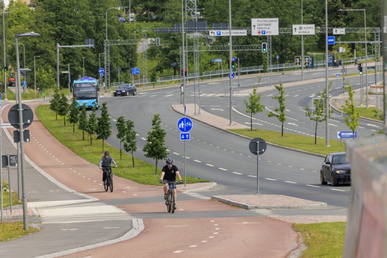 Kaksi pyöräilijää pyörätiellä vilkkaasti liikennöidyn Savilahdentien varrella.