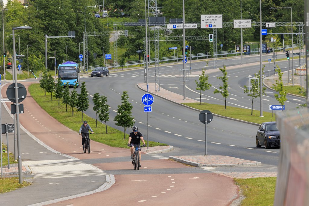 Kaksi pyöräilijää pyörätiellä vilkkaasti liikennöidyn Savilahdentien varrella.