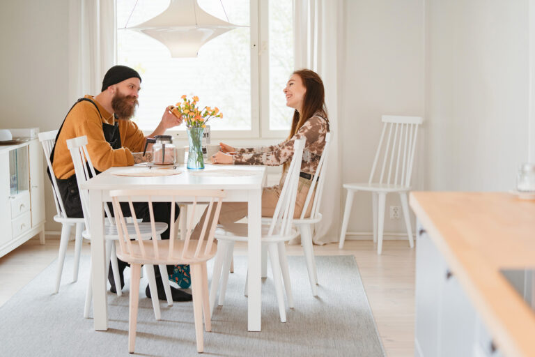 Nainen ja mies istuvat vaaleasävyisen keittiön pöydän ääressä, juovat kahvia ja nauravat.