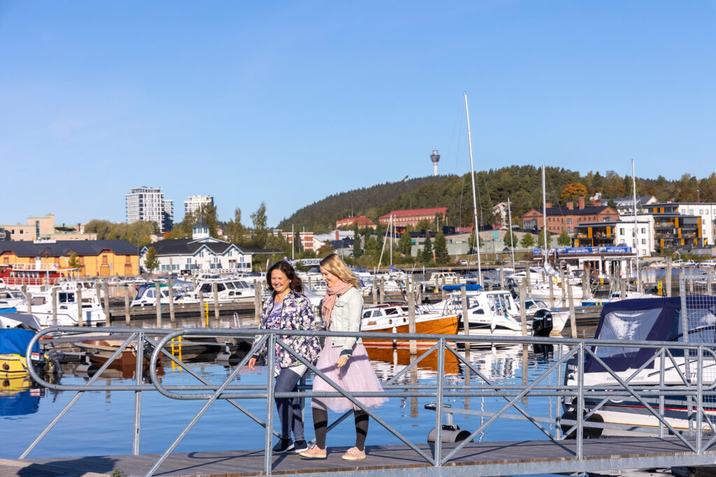 Kaksi naista kävelee sillalla Satamassa. Taustalla Kallavesi, veneitä ja Puijon torni.