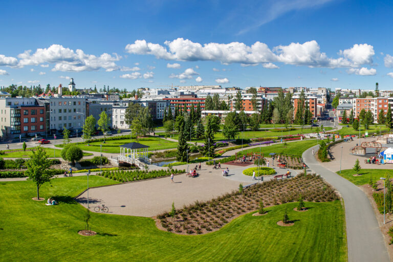 Vihreä puisto, jossa on paljon ihmisiä viettämässä kesäpäivää. Taustalla näkyy asuintaloja ja sininen taivas.