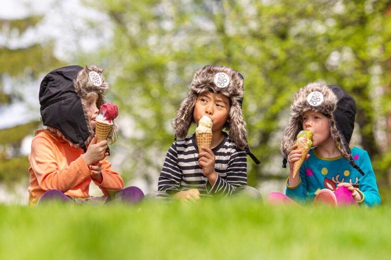 Kolme lasta istuu nurmikolla syömässä jäätelötötteröitä päässään karvahatut, joissa on I love Kuopio -pinssit.