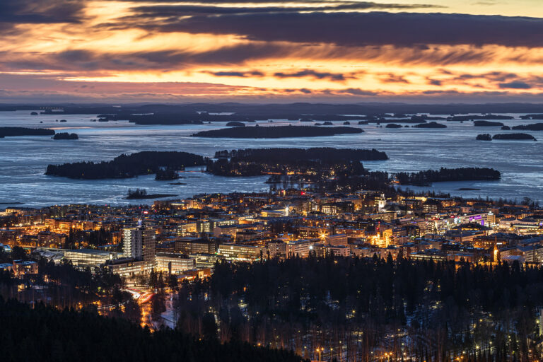 Kuopion maisema korkealta katsottuna auringonlaskun aikaan. Oranssi taivas, sininen järvi ja kaupunki upeana loistavissa valoissaan.