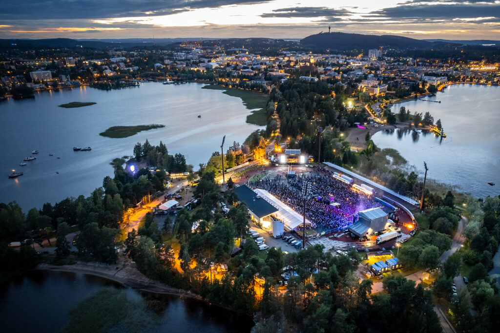 Kuopion keskusta-alue ilmasta katsottuna kesällä ilta-aikaan. Vettä, luontoa, Puijon torni ja ympäri kaupunkia hehkuvat valot. Etualalla Väinölänniemi, jossa on menossa konsertti ja ihmiset täyttävät koko stadionin alueen.