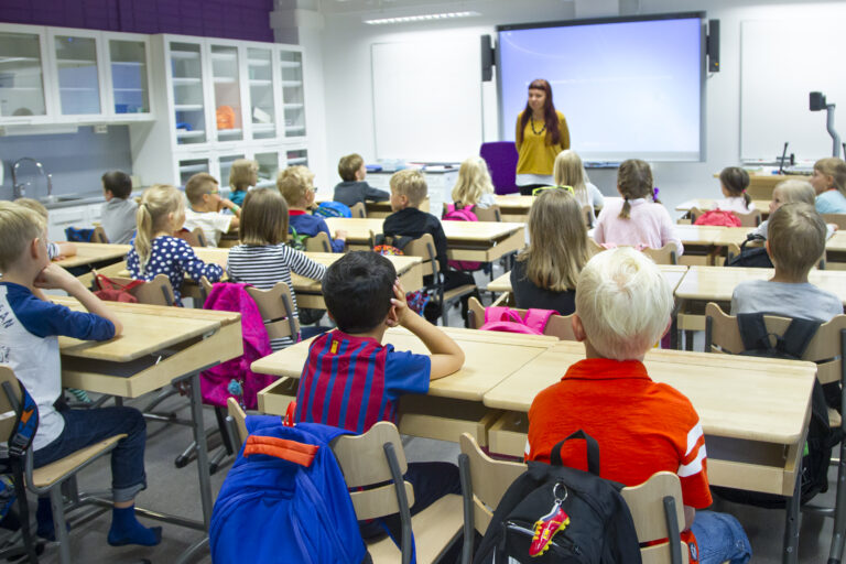 Oppilaita istumassa pulpeteissaan koululuokassa. Opettaja luokan edessä.