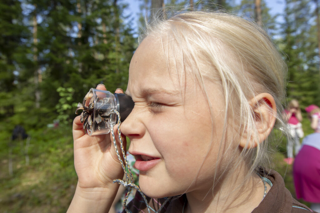 Alakouluikäinen oppilas oppimassa ulkona tutkien luupilla käpyä.