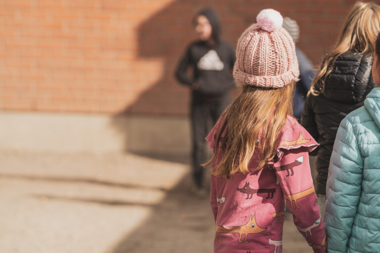 Alakouluikäinen lapsi (punertava pipo, pitkät hiukset ja punertava takki) etualalla kuvassa selkä kuvaajaan päin. Taustalla mustaan huppariin pukeutunut oppilas (epätarkkana kuvana).