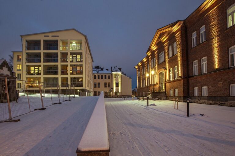 Kuva talvisesta kaupunkipihasta jonka taustalla on lämpimästi valaistuja kaupunkikerrostaloja