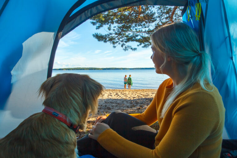 Vaaleahiuksinen nainen ja koira istuvat teltassa rannalla ja katsovat järvellepäin. Rantaviivassa seisoo kaksi lasta värikkäät pyyhkeet päällään. On kesä ja aurinko paistaa.
