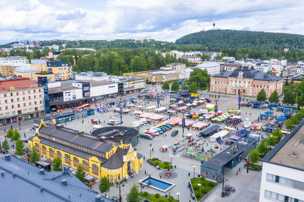 Kuopion kauppahalli, kaupungintalo ja Puijon torni sekä tori täynnä myyntikojuja ja ihmisiä.
