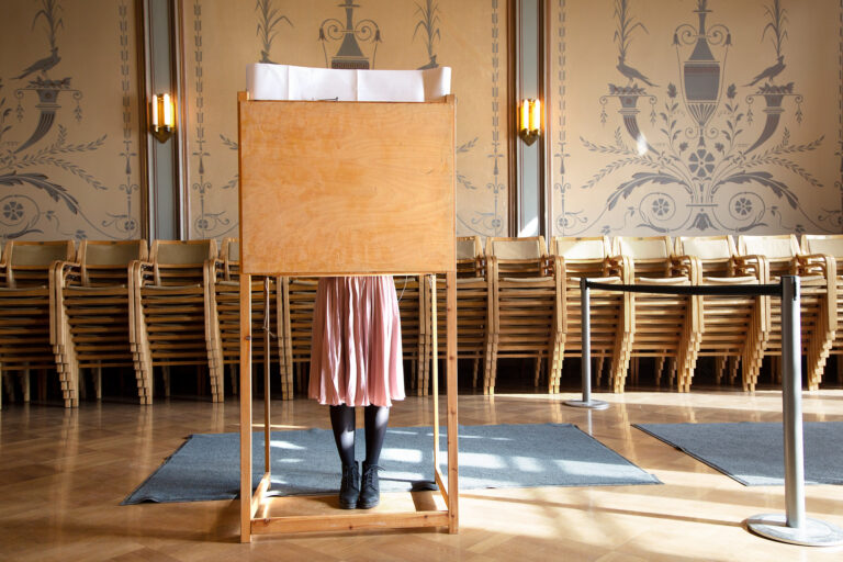 Nainen äänestämässä puisessa äänestyskopissa vaaleanpunainen hame päällä.